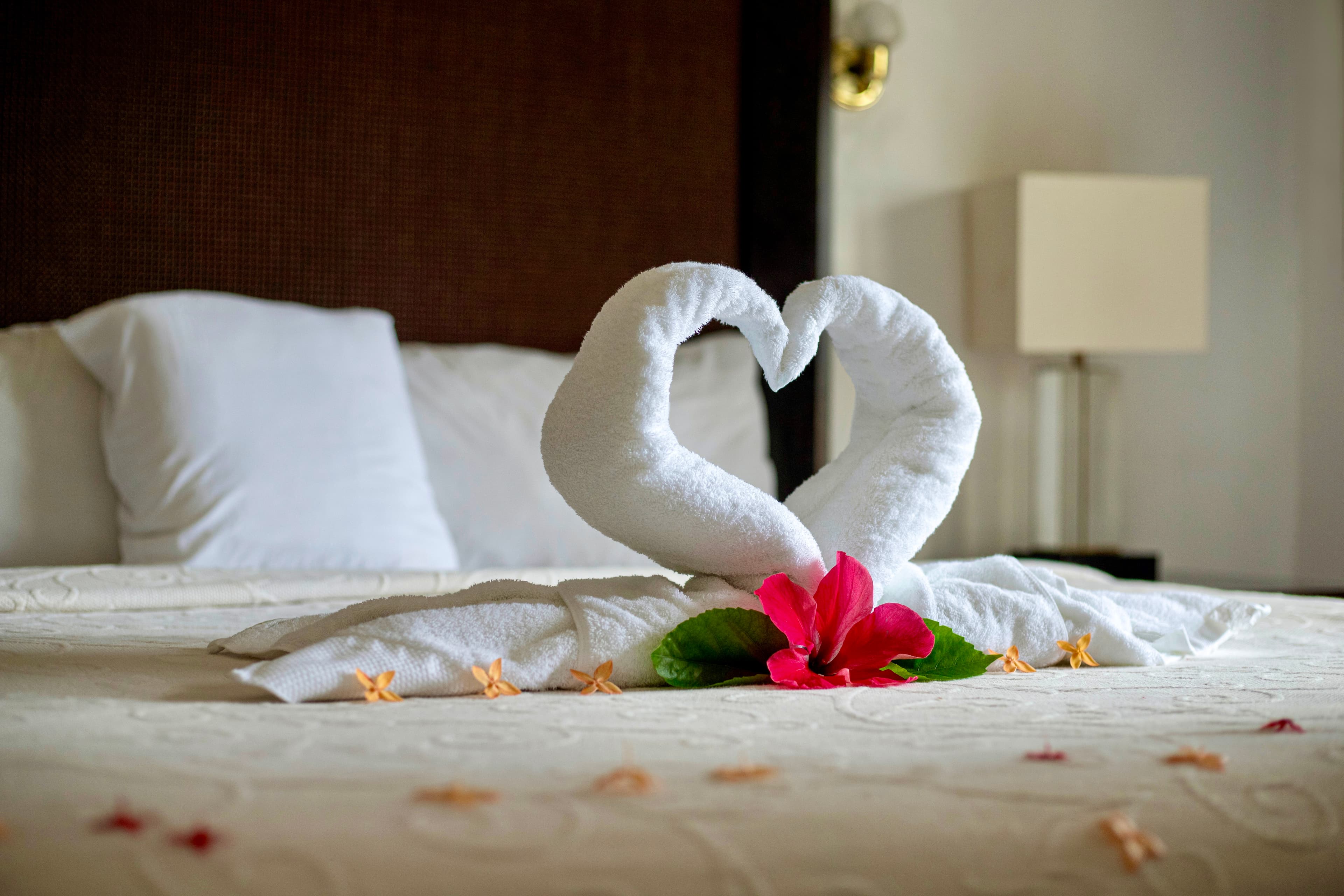 A beautifully made bed with white linens features two towels folded into a heart shape, adorned with a red hibiscus.