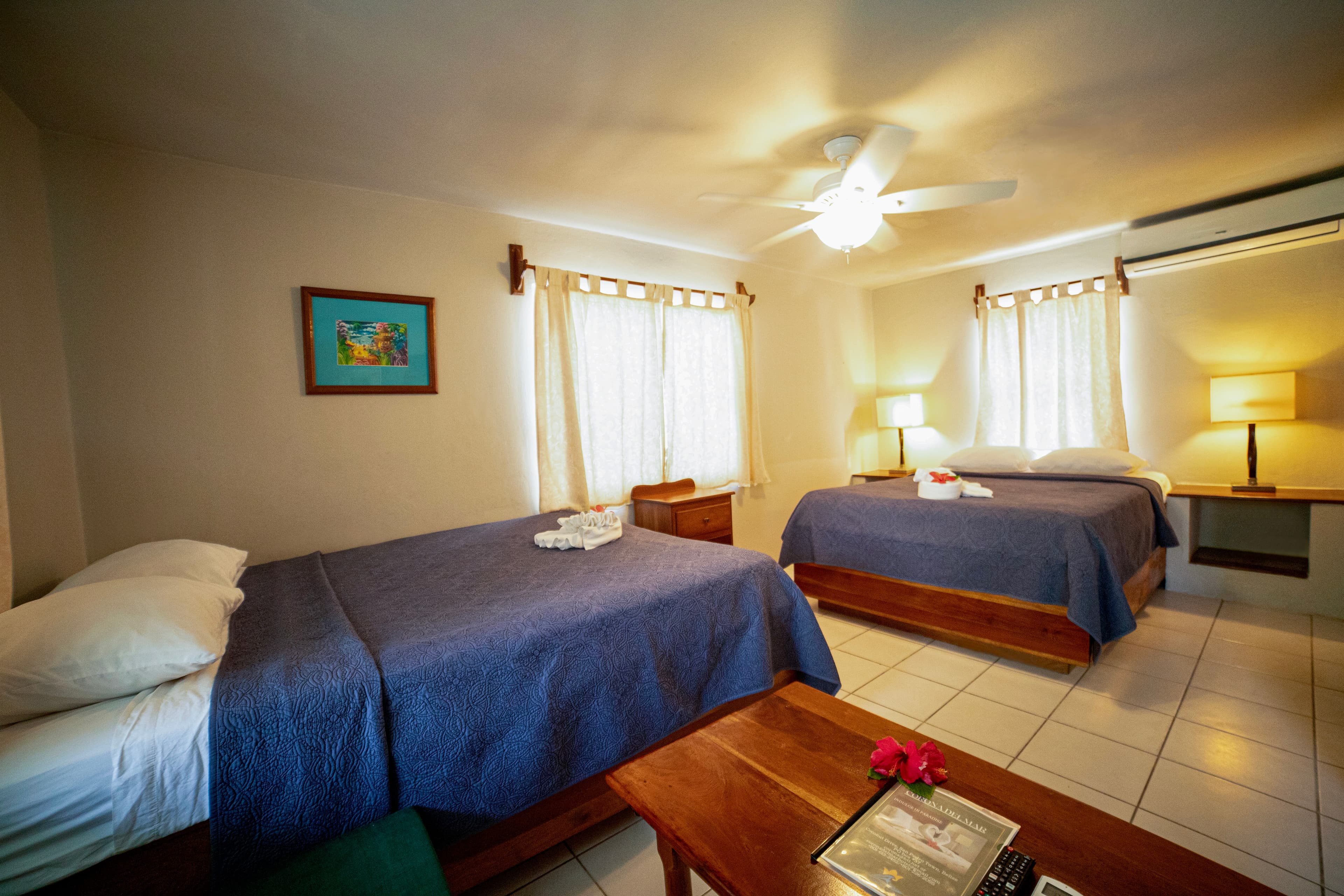 A bright hotel room featuring two queen beds with blue covers, a ceiling fan, and air conditioning, ready for a comfortable stay.