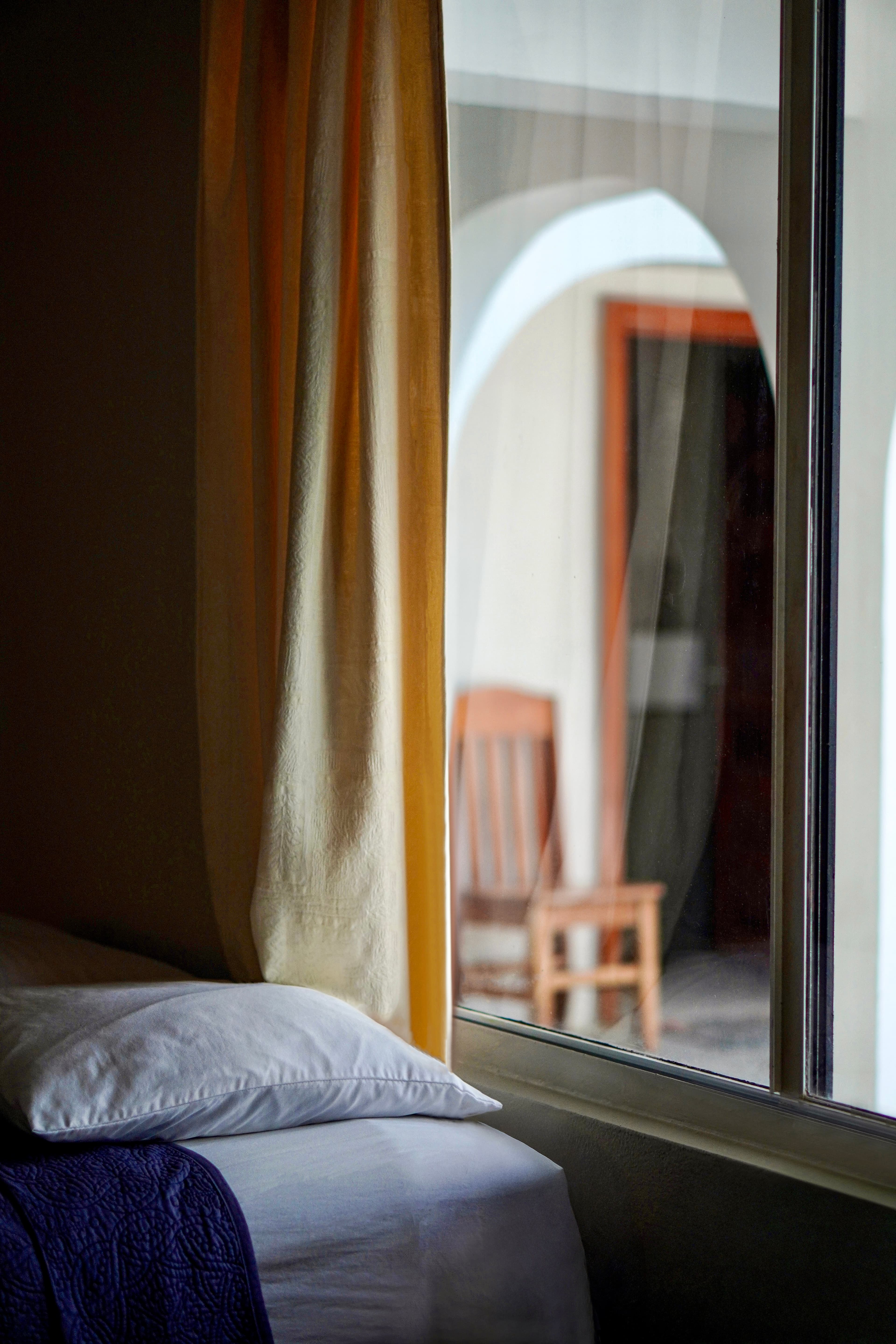 A cozy bed with white linens and a purple throw blanket, seen through a window with a sheer curtain, overlooking an arched patio.