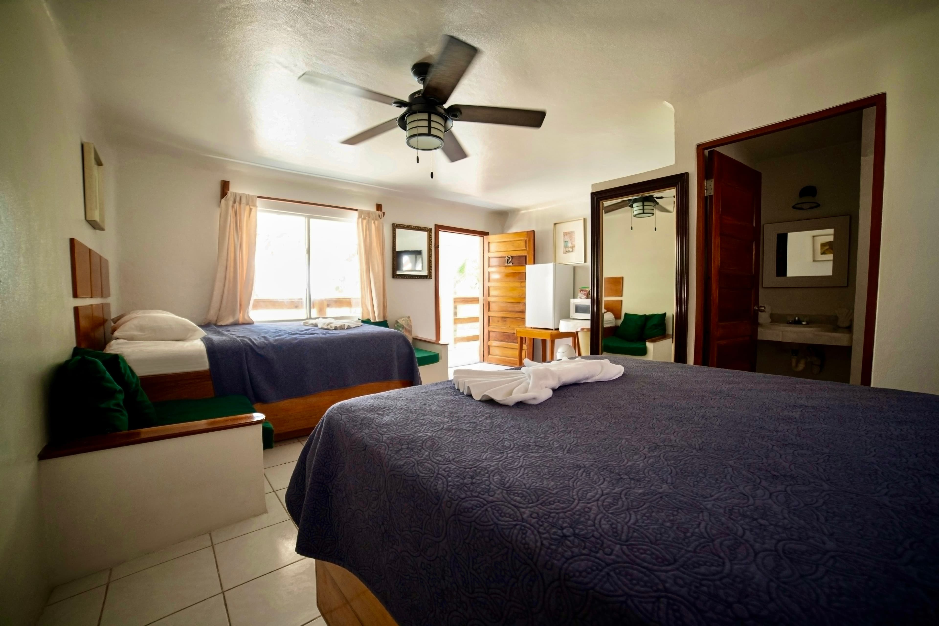 A spacious hotel room with two queen beds, blue blankets, a ceiling fan, and a view into the bathroom