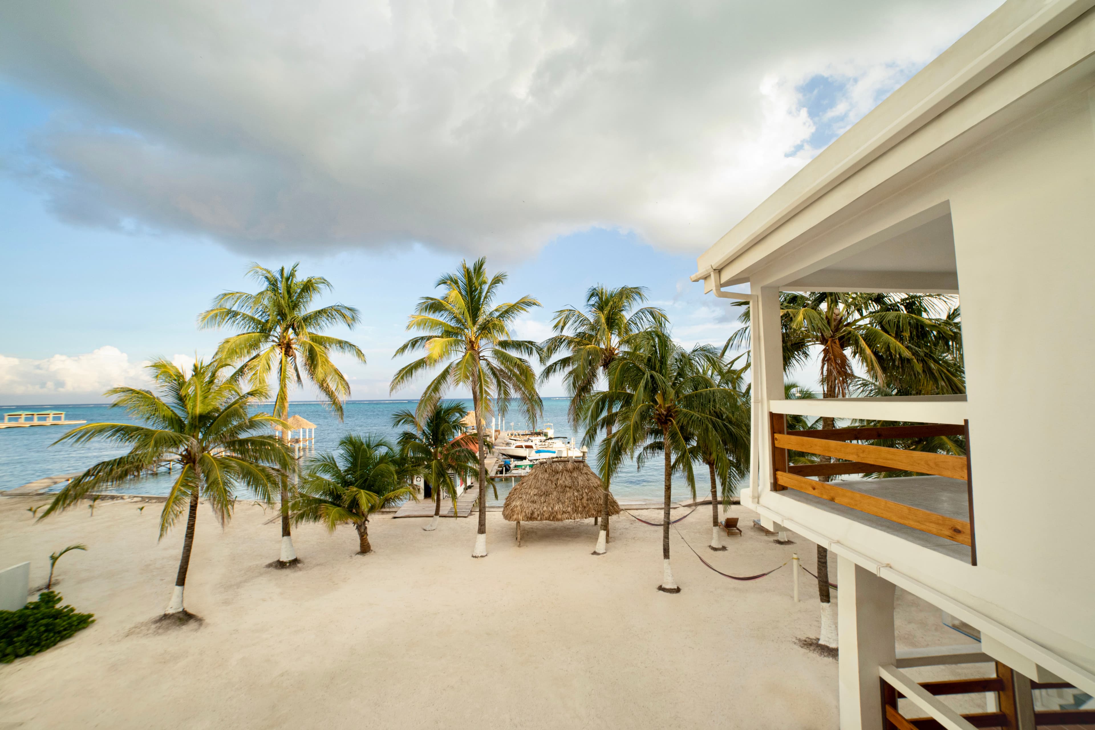 A scenic view of a white sandy beach with swaying palm trees, a thatched hut, and the ocean under a cloudy sky, from a building balcony.