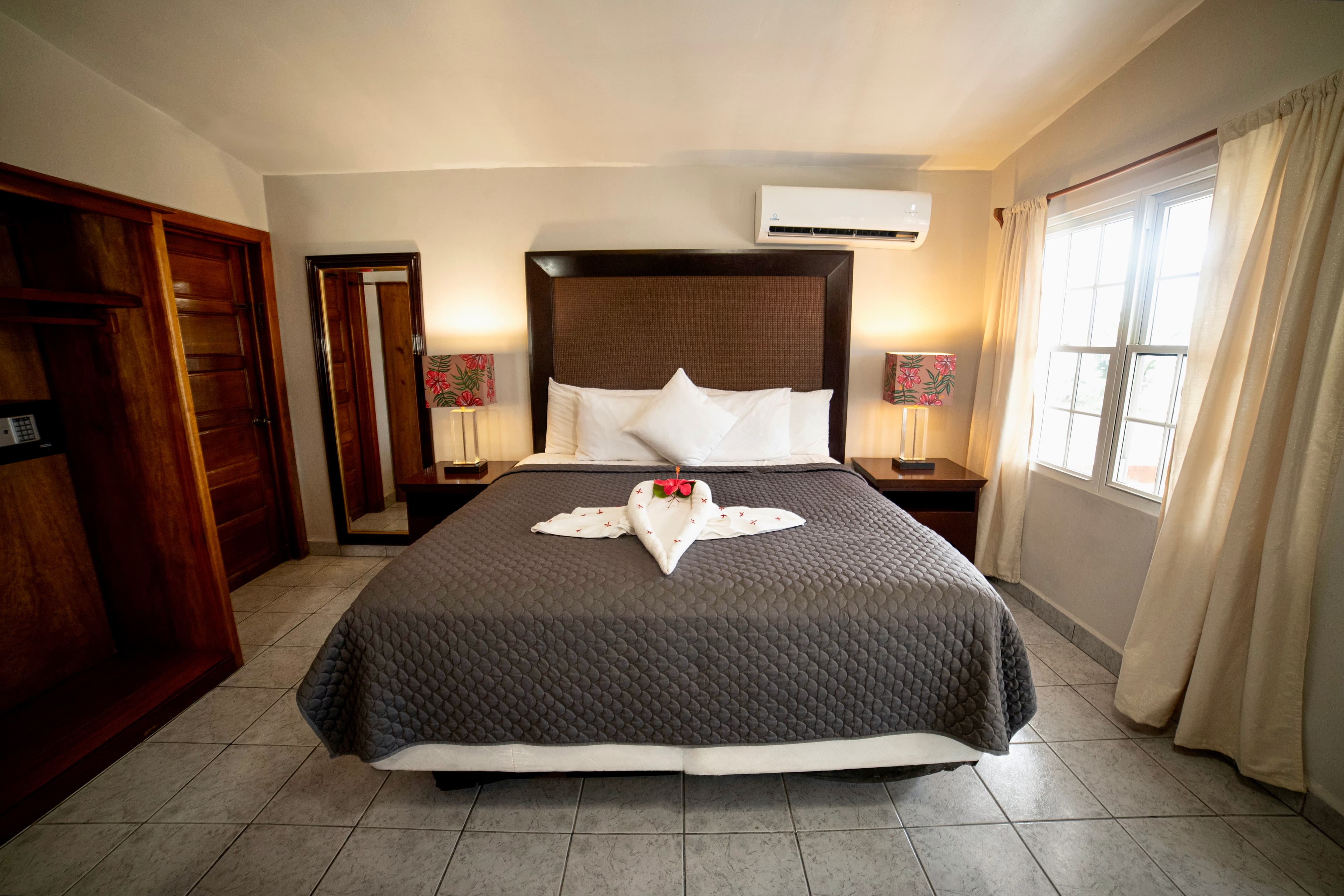 A spacious hotel room features a bed with a dark gray quilt, white folded towels, two bedside lamps, and an air conditioning unit.