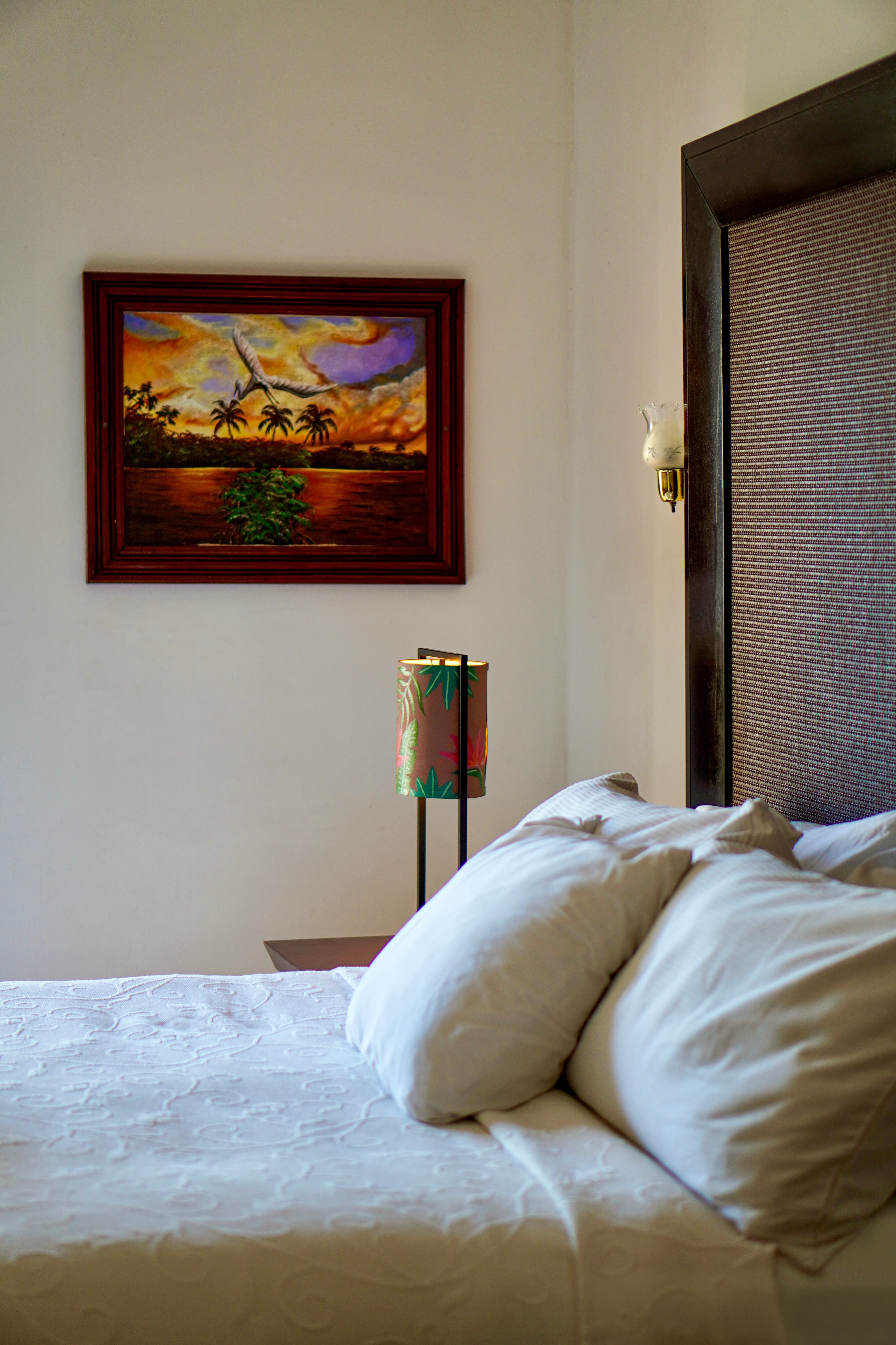 A comfortable bed with white pillows and sheets, a unique lamp, and a vibrant painting of a landscape with palm trees on the wall.