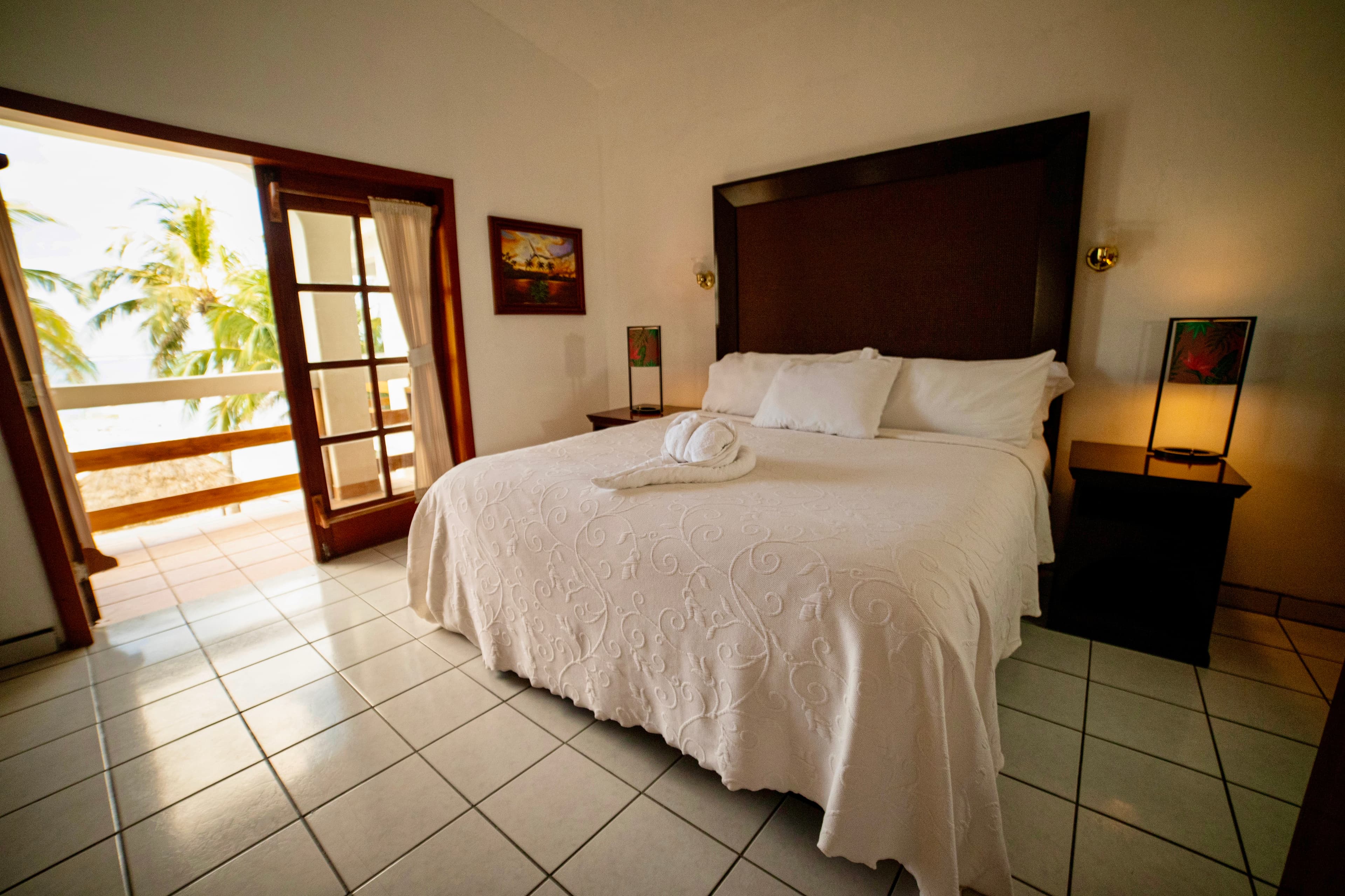 A serene bedroom with a large bed, white linens, and a private balcony overlooking a beach with palm trees and the ocean.