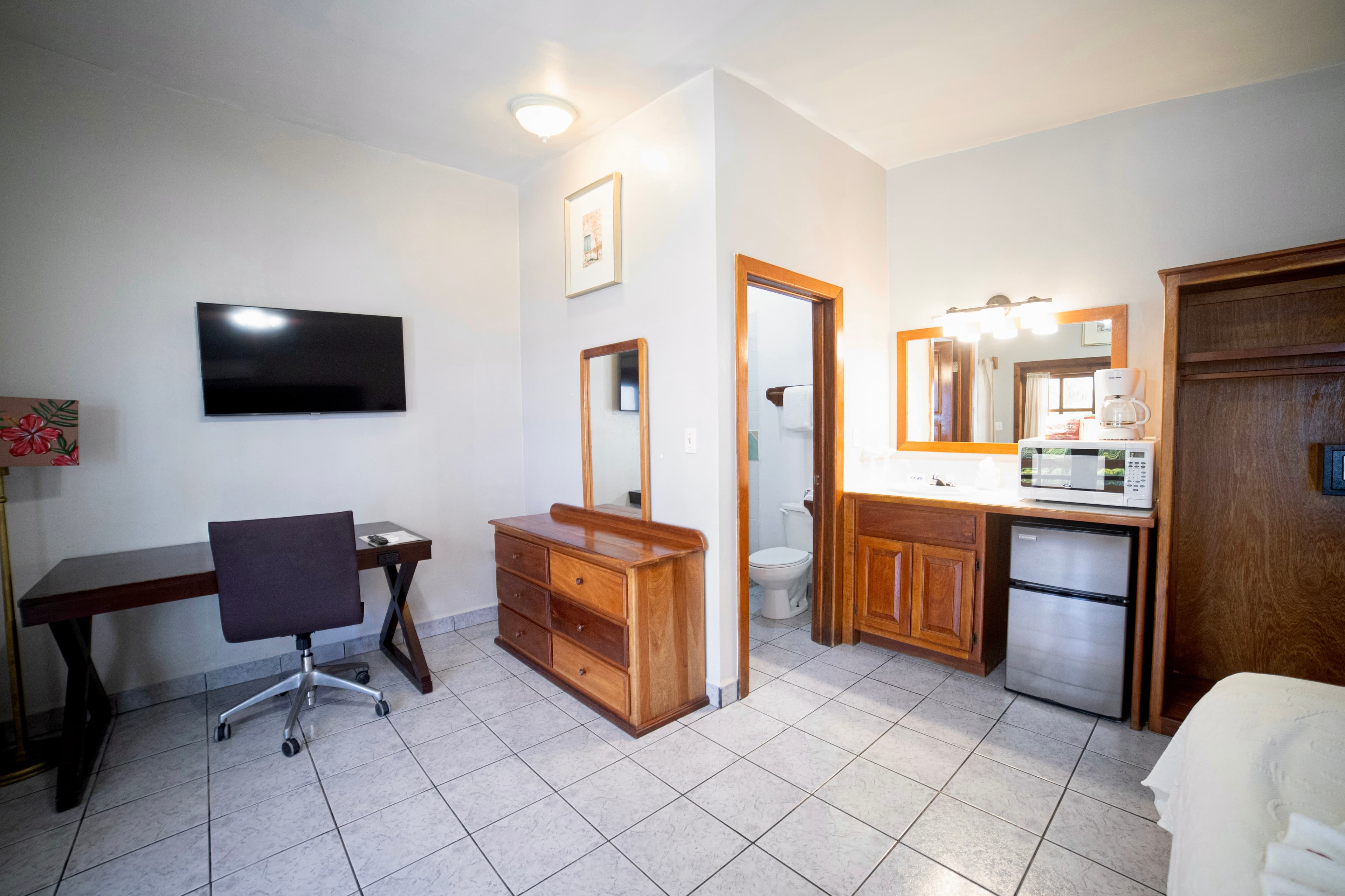 A hotel room featuring a desk with an office chair, a wall-mounted TV, a dresser, and a kitchenette area with a microwave and mini-fridge.