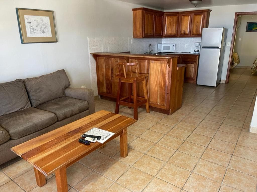An inviting hotel room featuring a brown sofa, a wooden coffee table, a bar-style dining area, and a functional kitchen with appliances.