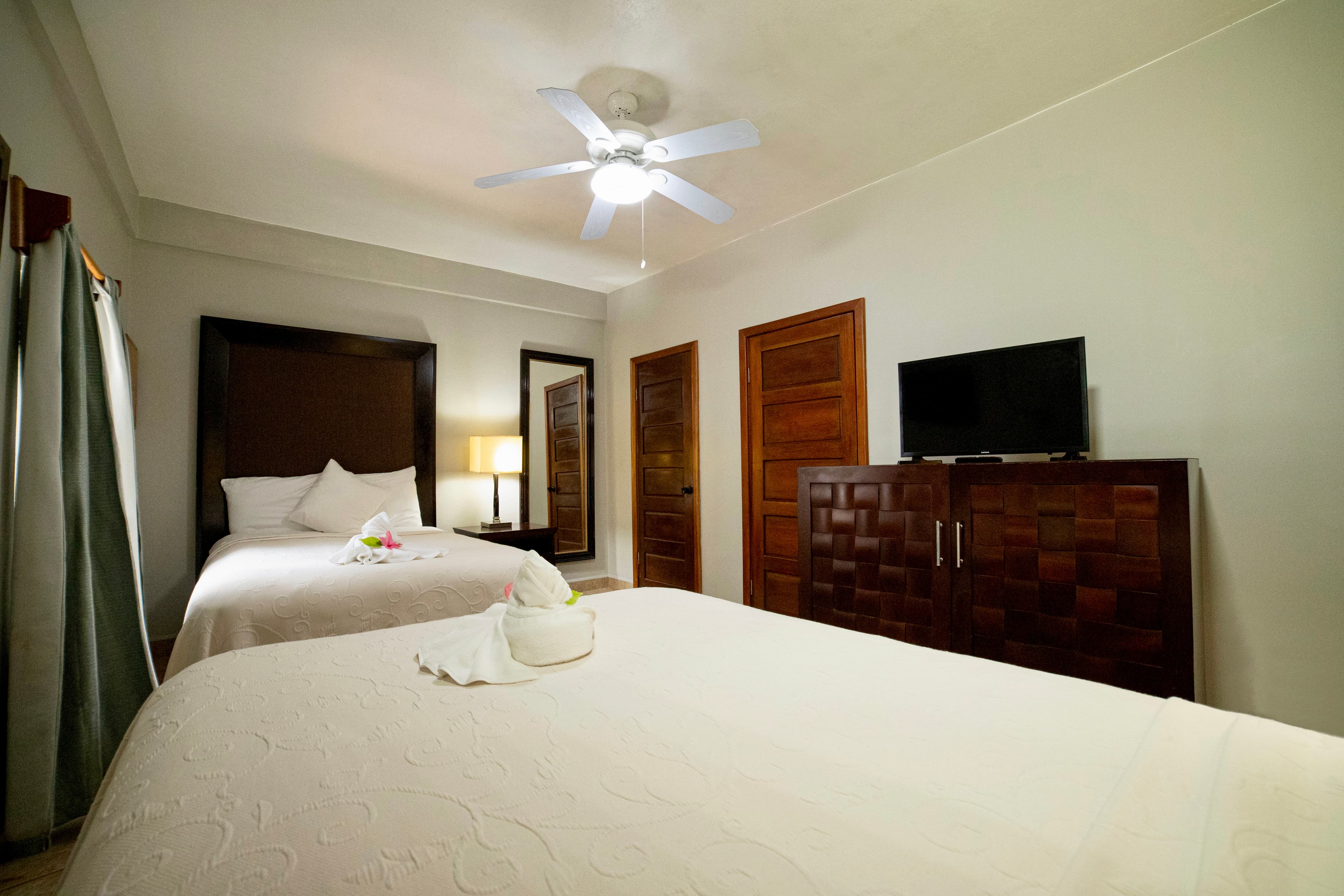 A comfortable hotel room with two beds, white linens, a ceiling fan, a dark wooden dresser with a TV, and a full-length mirror.