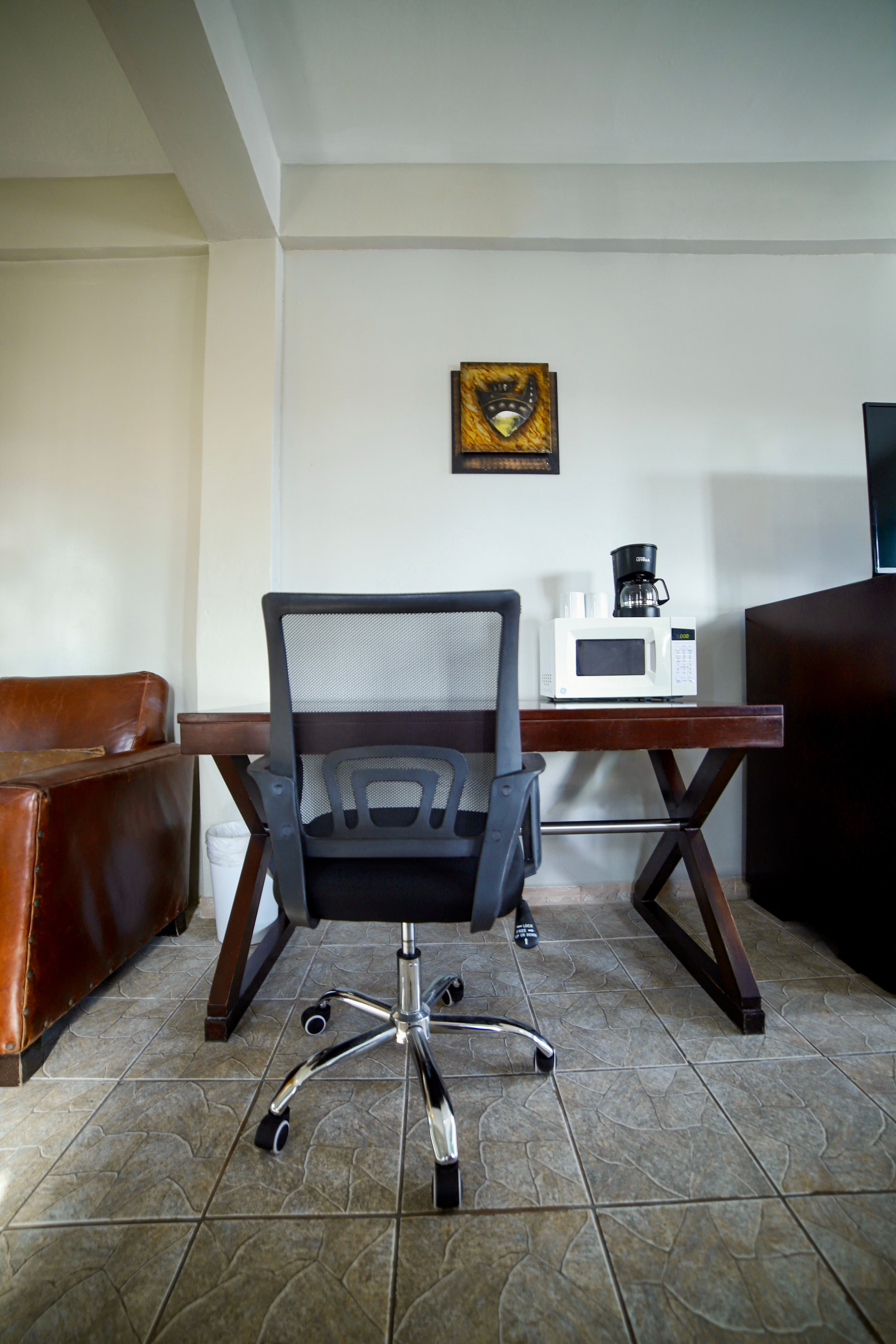 A minimalist workspace featuring a dark wooden desk, a black mesh office chair, a microwave, and a coffee maker, set against a white wall.