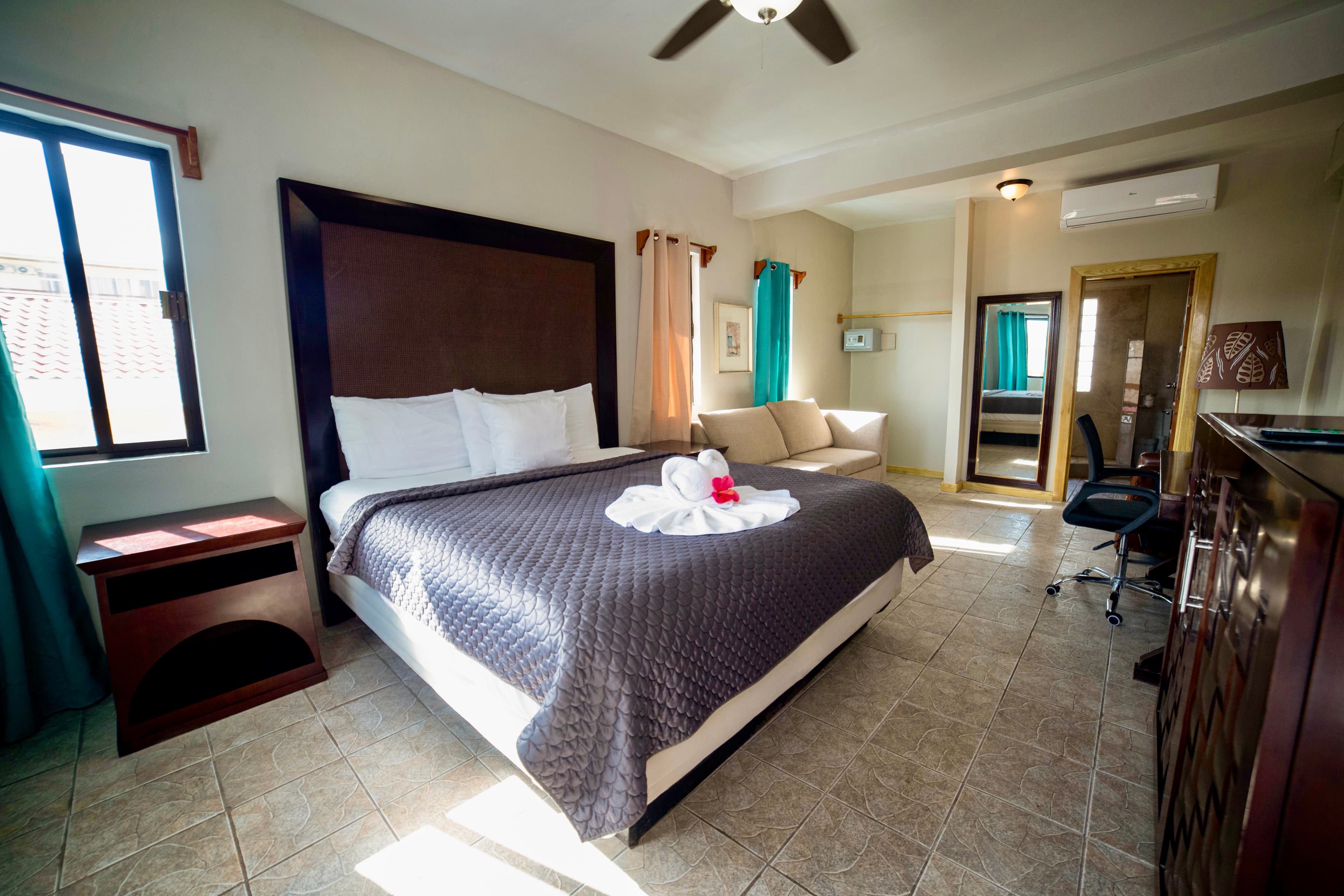 A bright hotel room with a large bed, dark gray quilt, white towels, a ceiling fan, and a light-colored couch.