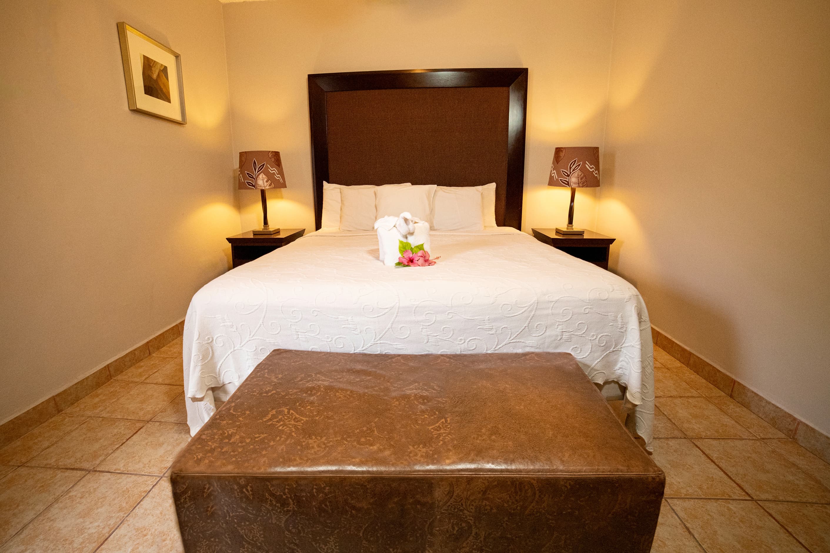 A luxurious bedroom features a king-size bed with white linens, two bedside lamps casting a warm glow, and a brown ottoman at the foot of the bed.