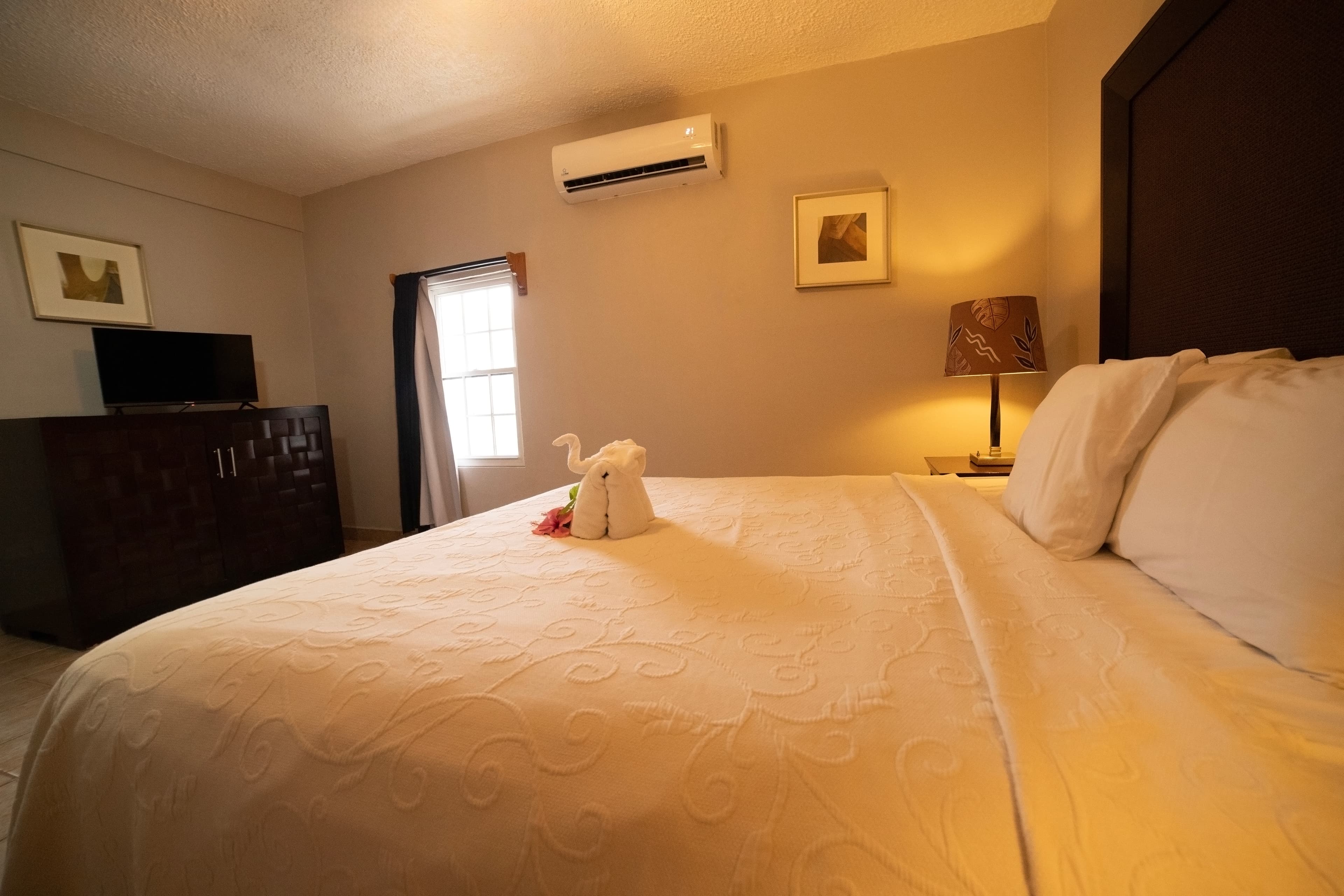 A spacious and inviting hotel room with a large bed, textured white bedding, a creative towel animal, and a wall-mounted air conditioner.