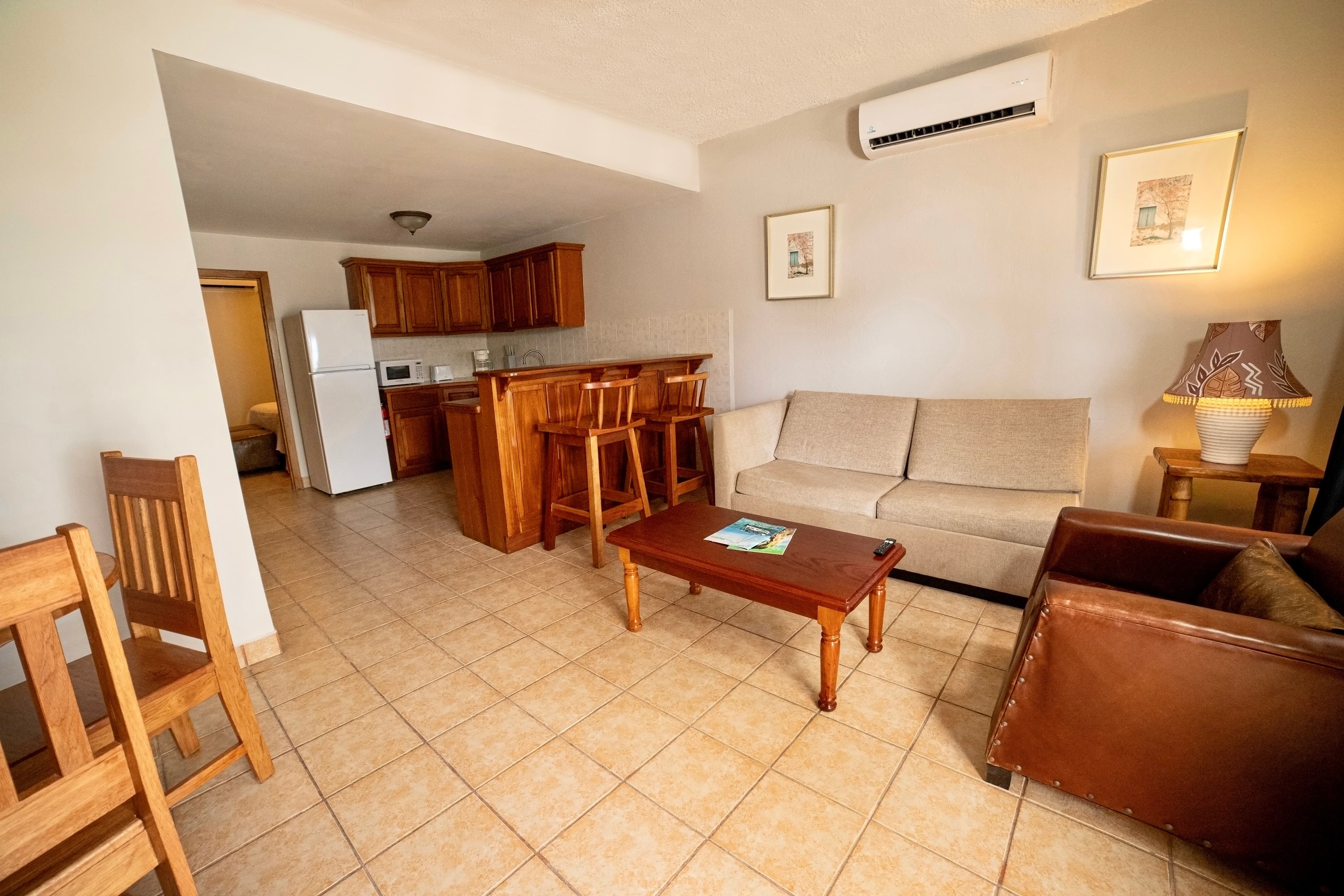 A bright, open-plan living area with a beige sofa, brown armchairs, a wooden coffee table, a kitchen with a breakfast bar, and a dining area.