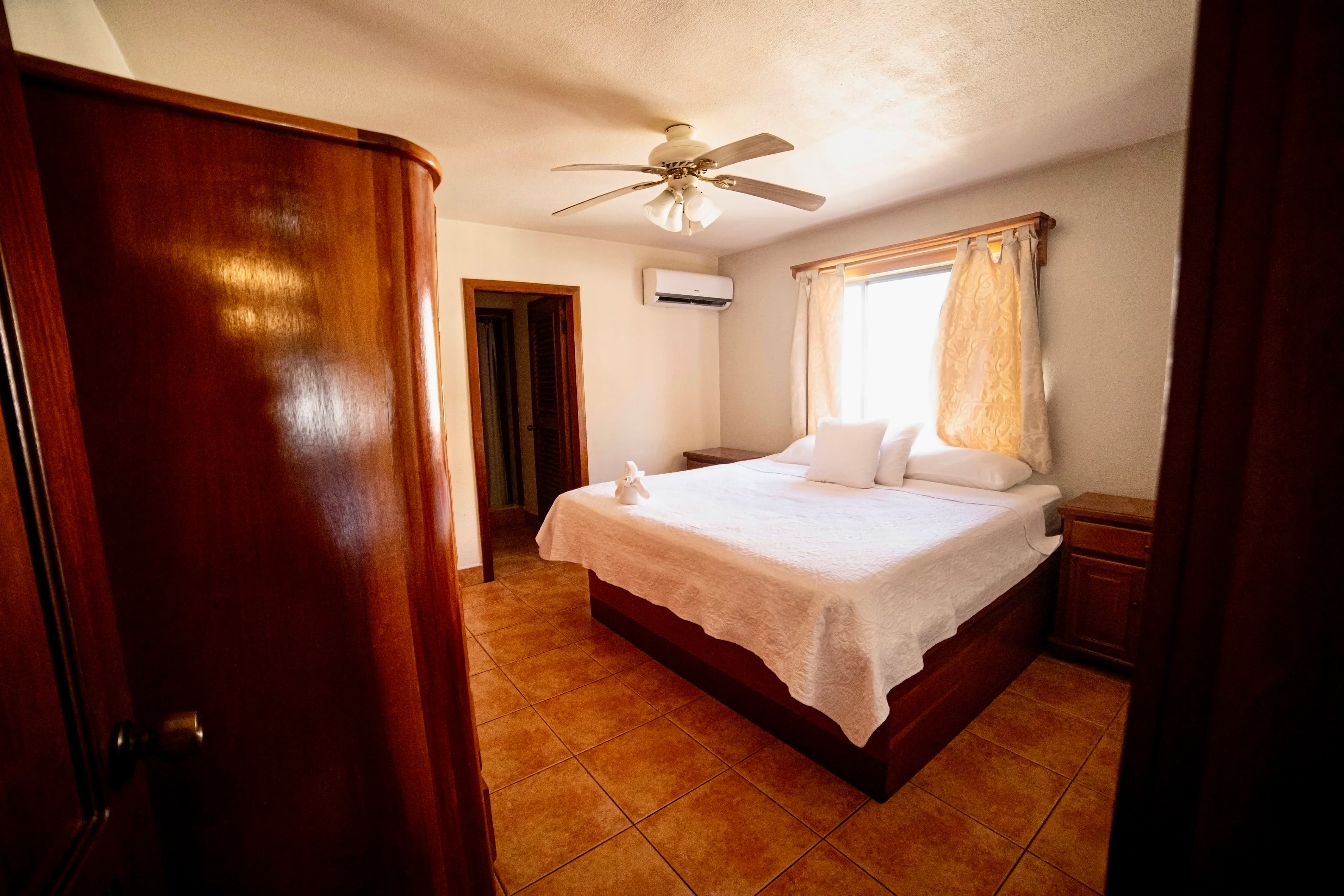 A well-lit bedroom with a large bed, white linens, a ceiling fan, and an air conditioning unit.