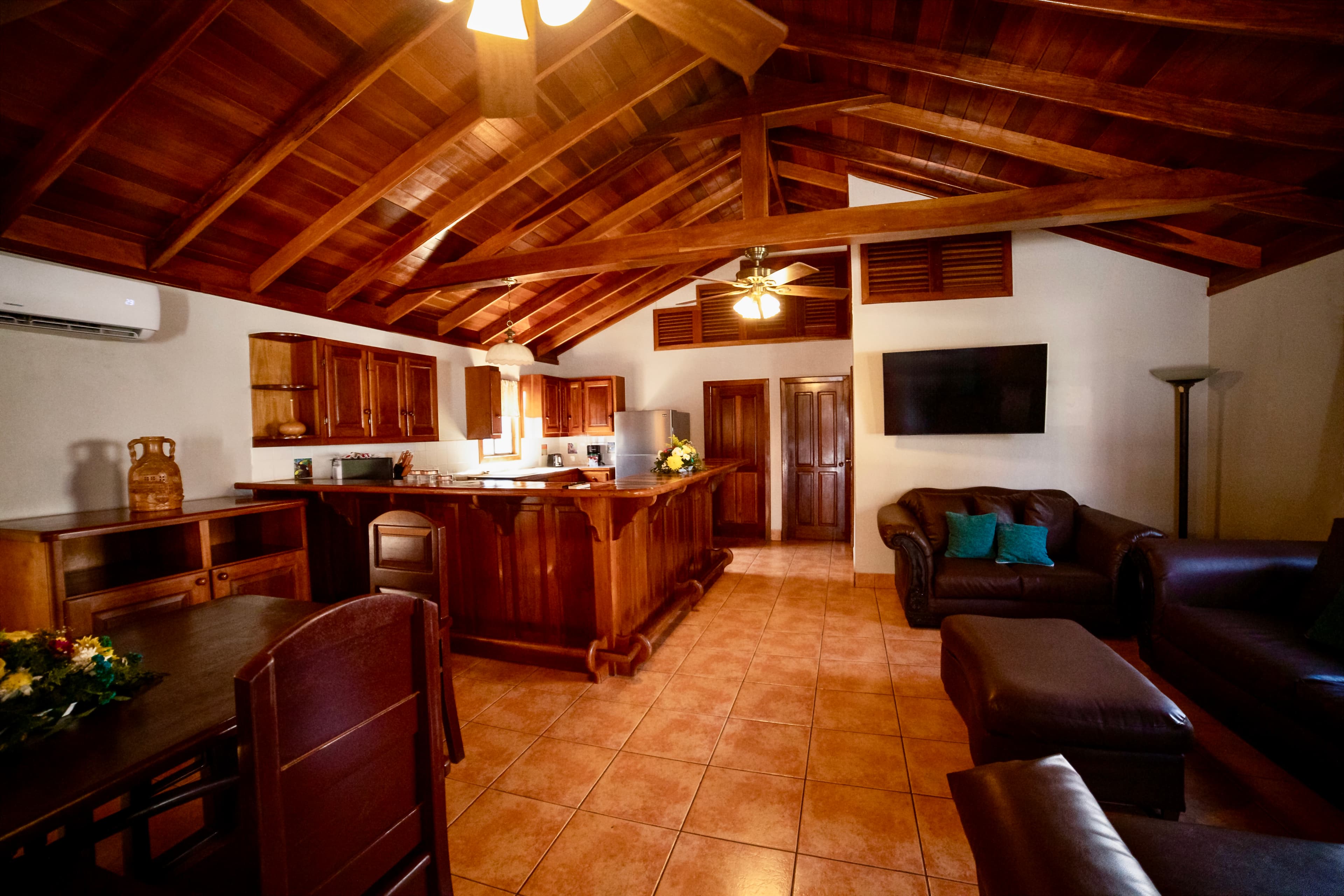 A spacious living area with vaulted wooden ceilings, a full kitchen with a bar, a comfortable seating area with dark sofas, and a dining space.