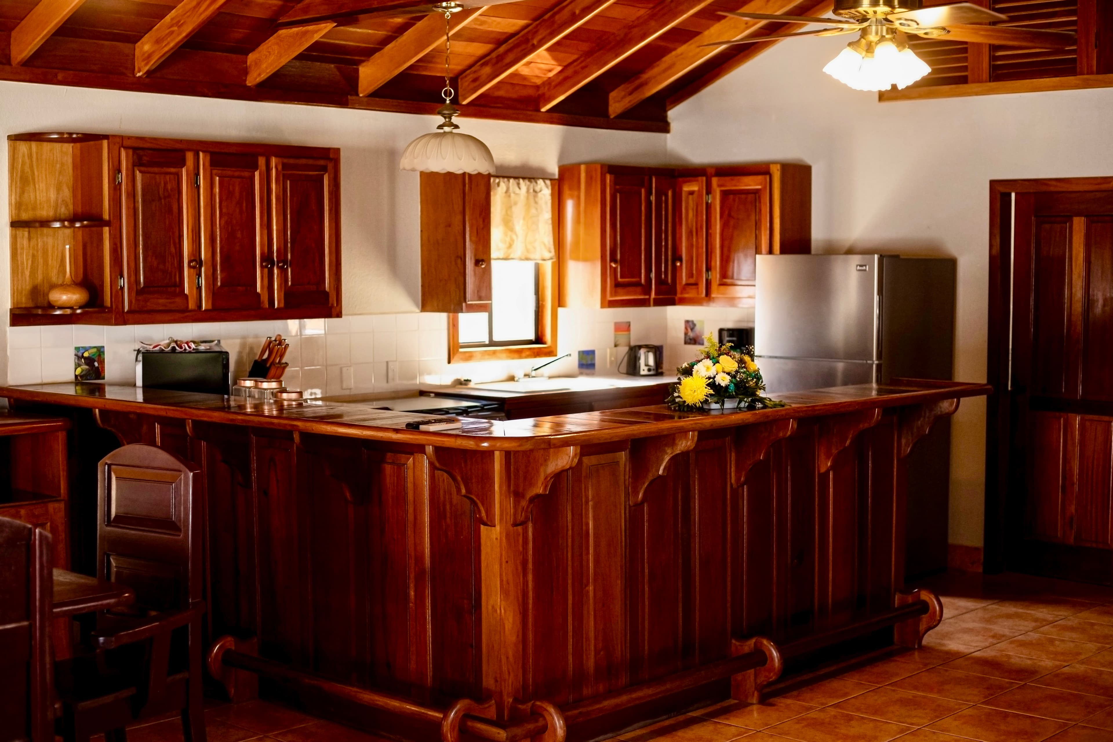 A rustic kitchen features a polished wooden bar and cabinets, complete with a sink, microwave, coffee maker, and a stainless steel fridge.