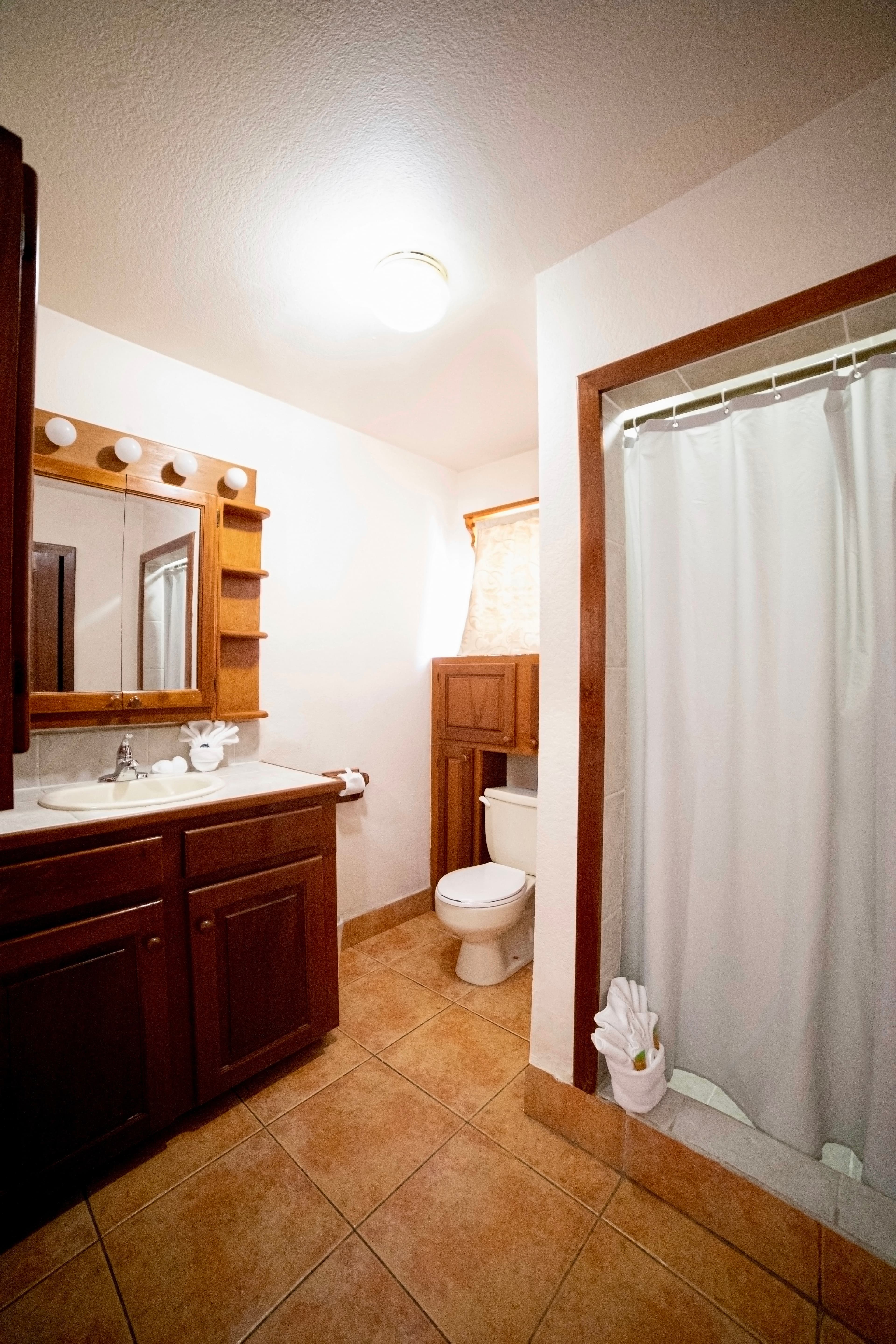 A clean bathroom with tiled floors, a dark wood vanity with a sink and mirror, a toilet, and a walk-in shower with a white curtain.