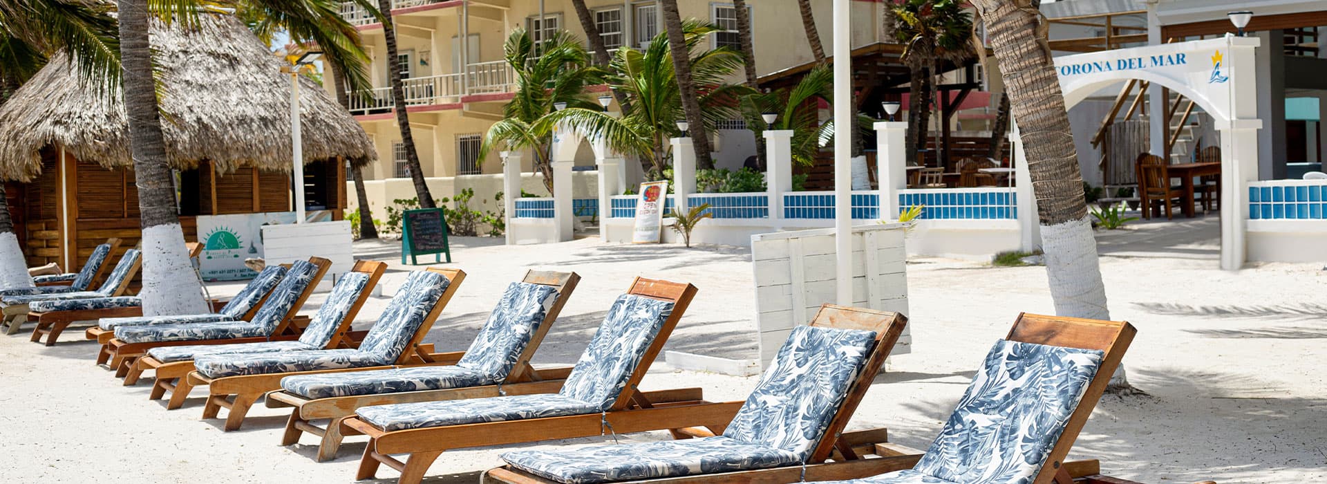 Beach loungers with patterned cushions lined up on a sandy shore near palm trees and a thatched roof structure.
