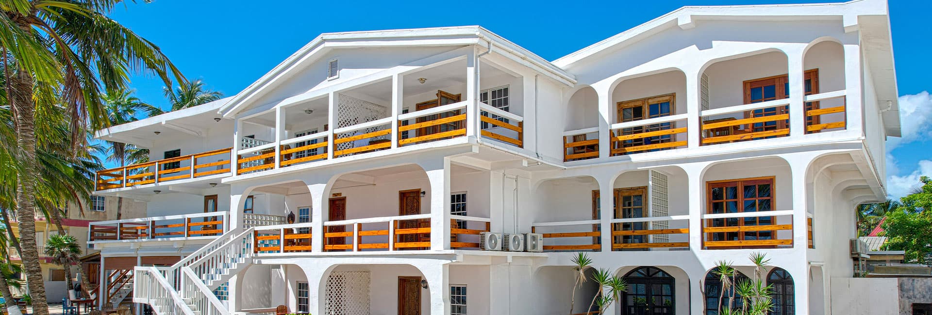 A white multi-story building with wooden balconies surrounded by palm trees against a clear blue sky.