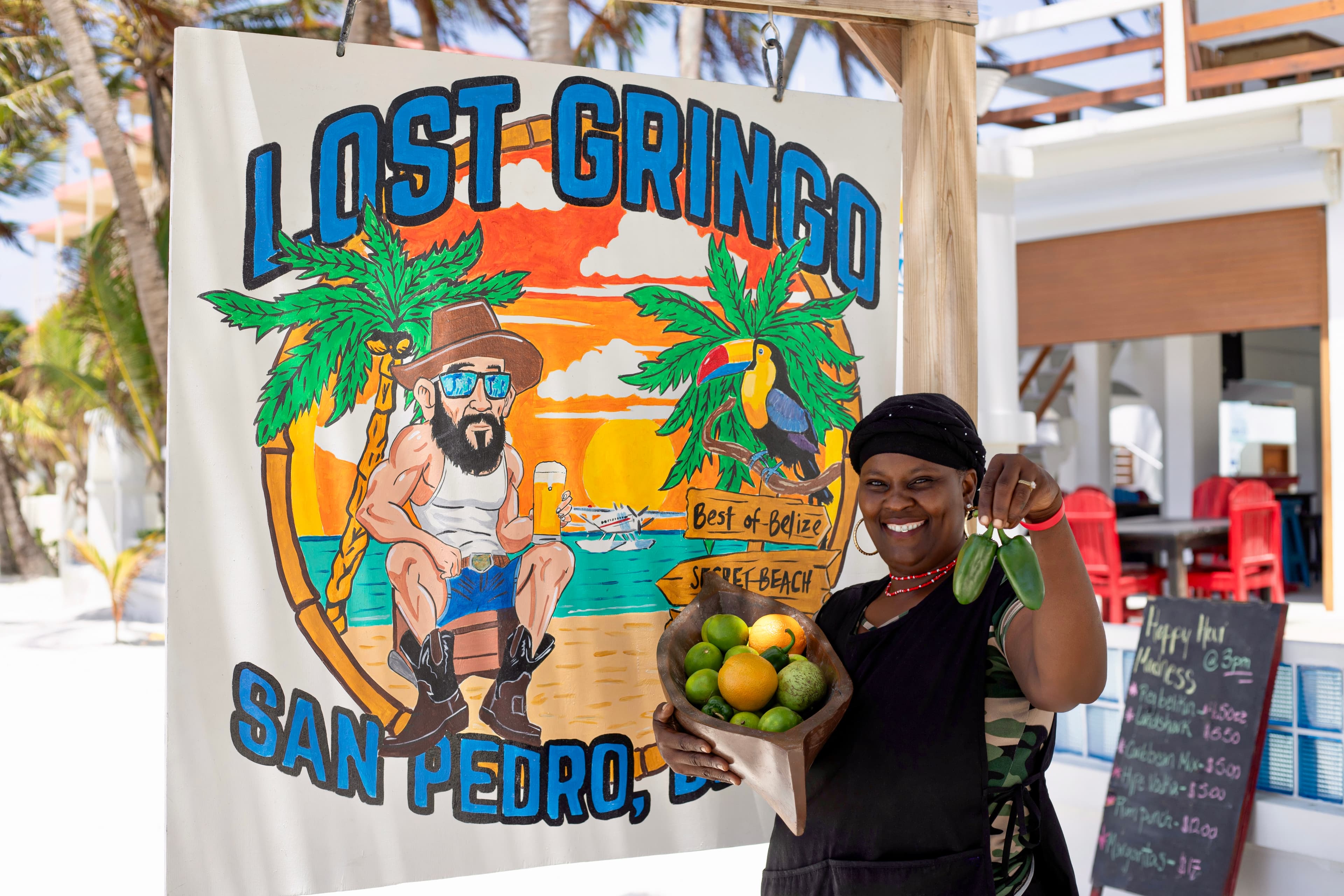 A smiling woman holds a bowl of fruits and vegetables in front of a colorful sign for "Lost Gringo" in San Pedro, Belize.