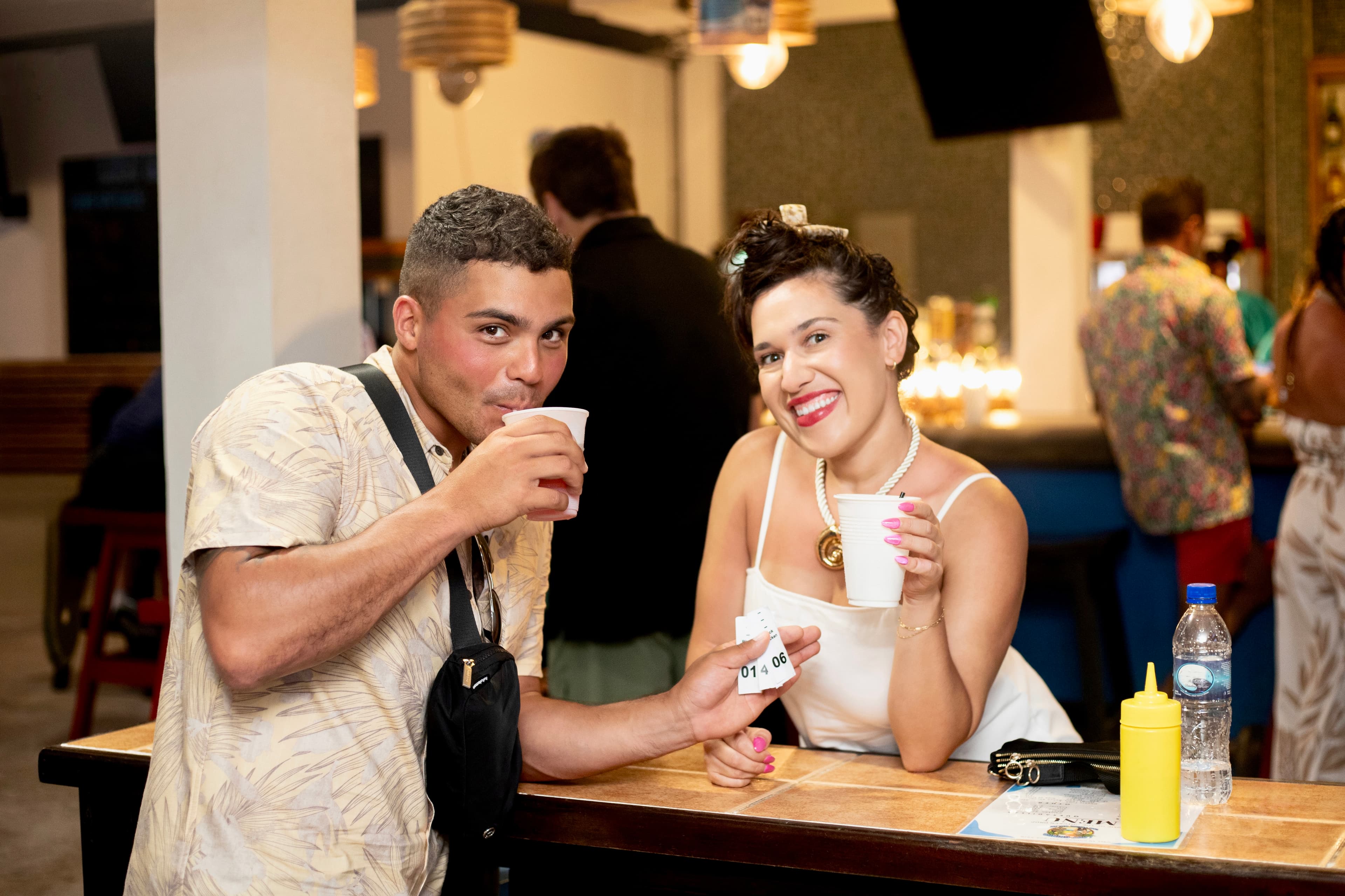 Two smiling friends hold cups at a bar while posing for the camera.