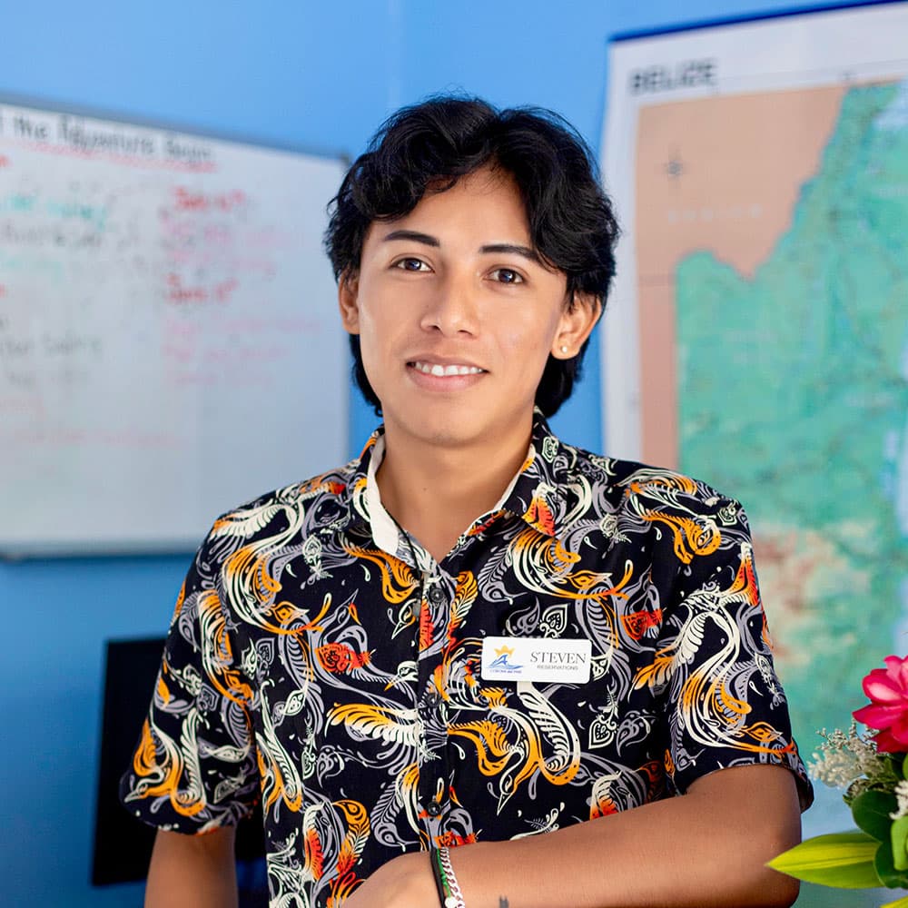 A young man with dark, curly hair stands smiling in a colorful shirt, in front of a blue wall and a map.