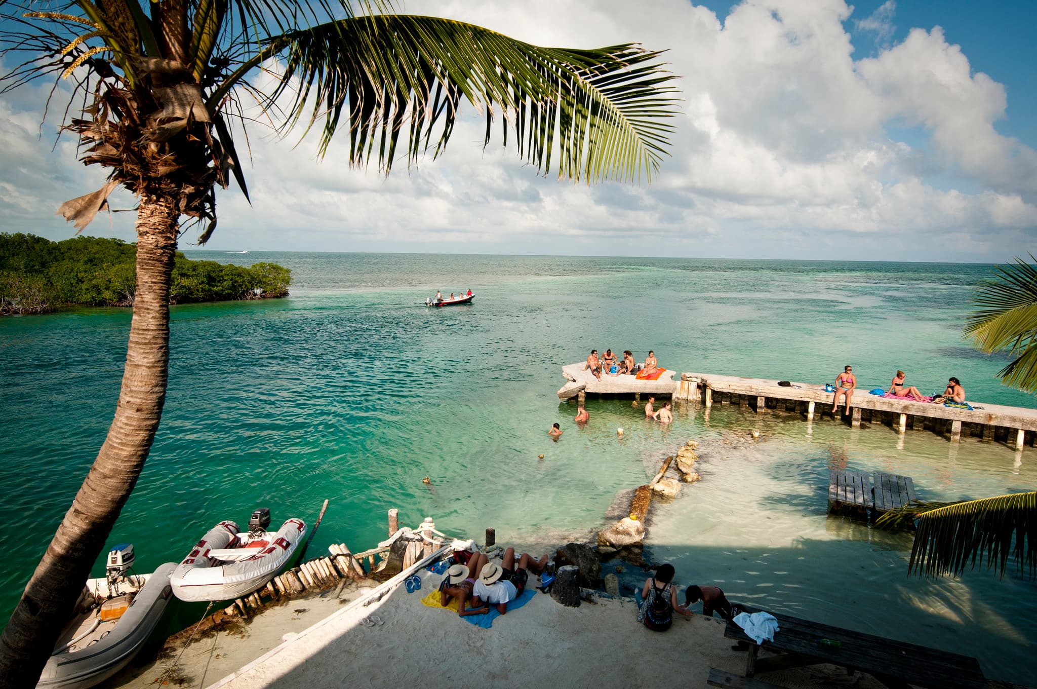 Tropical scene featuring people relaxing by turquoise waters, a dock, and a boat in the distance under a partly cloudy sky.