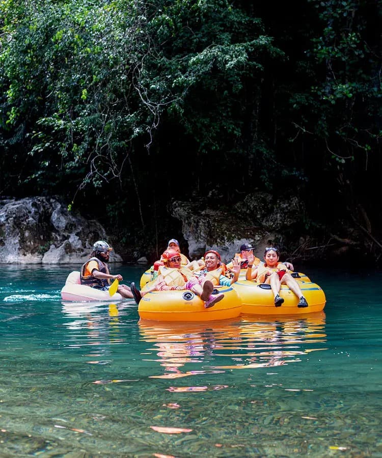 A group of six people in bright orange life jackets float on inflatable tubes in a calm, greenish-blue river surrounded by lush trees.