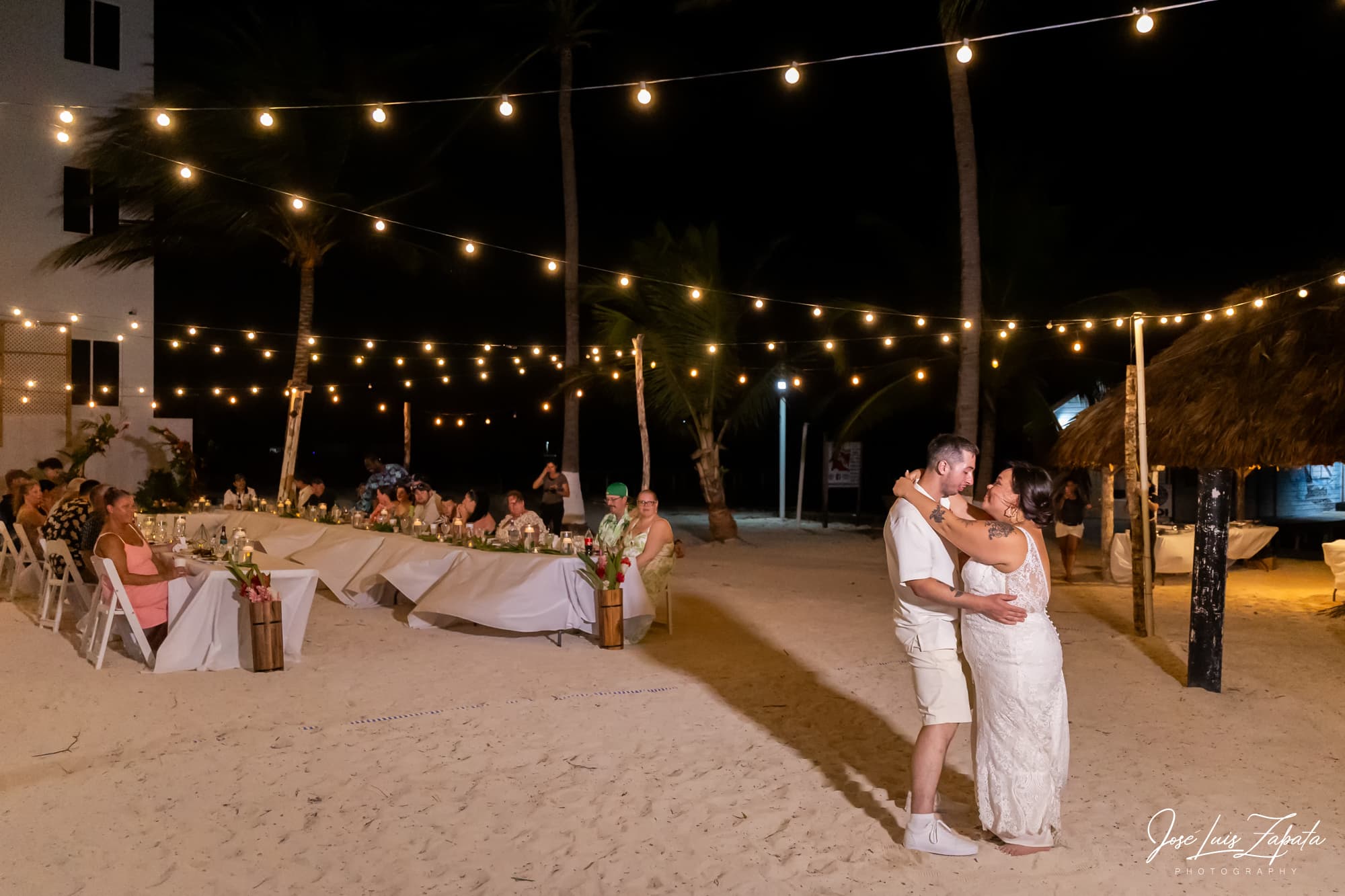 A couple dances under string lights at a beachfront wedding reception with guests seated at elegantly arranged tables.