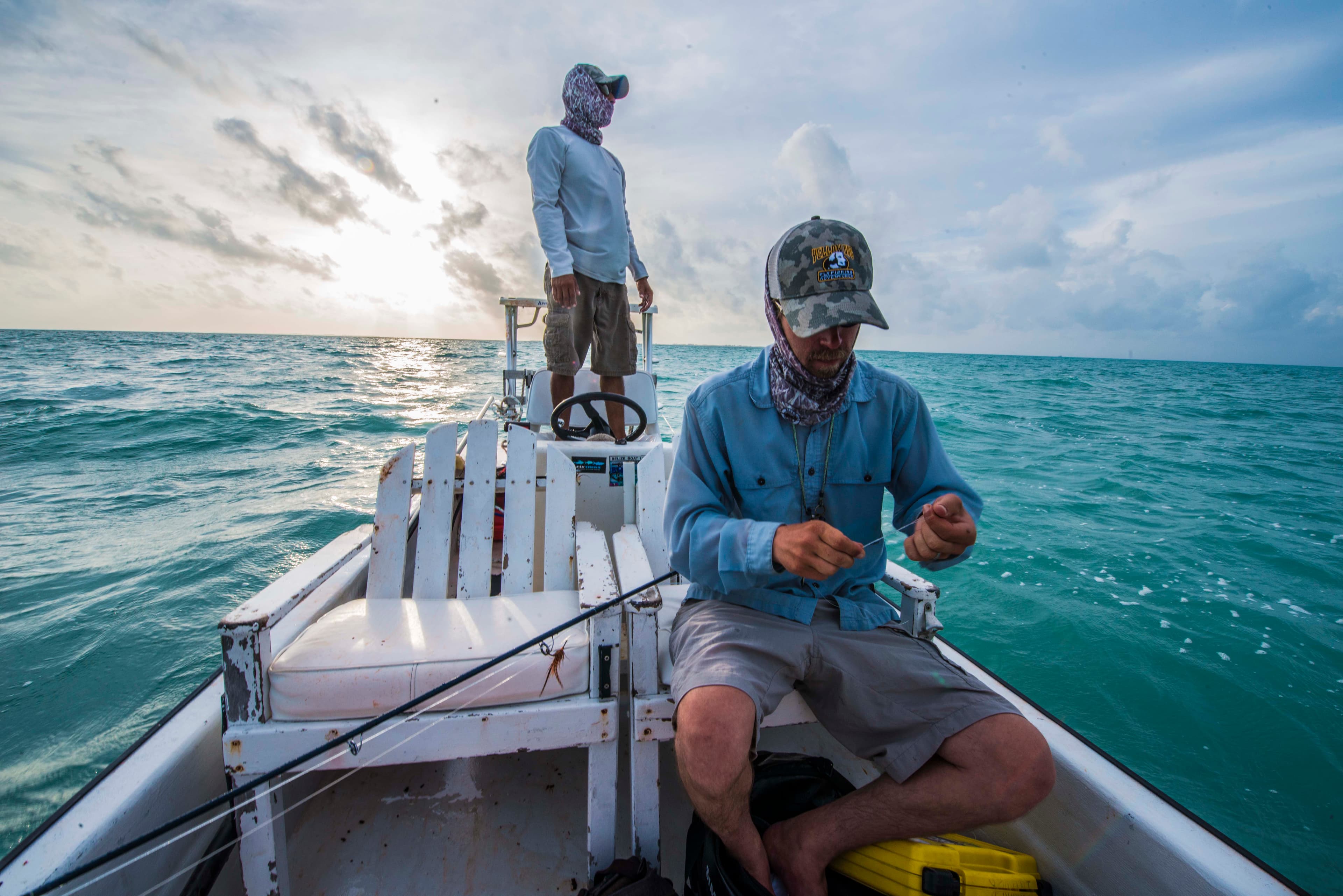Two fishermen on a boat, with one preparing bait as the sun sets over the ocean.