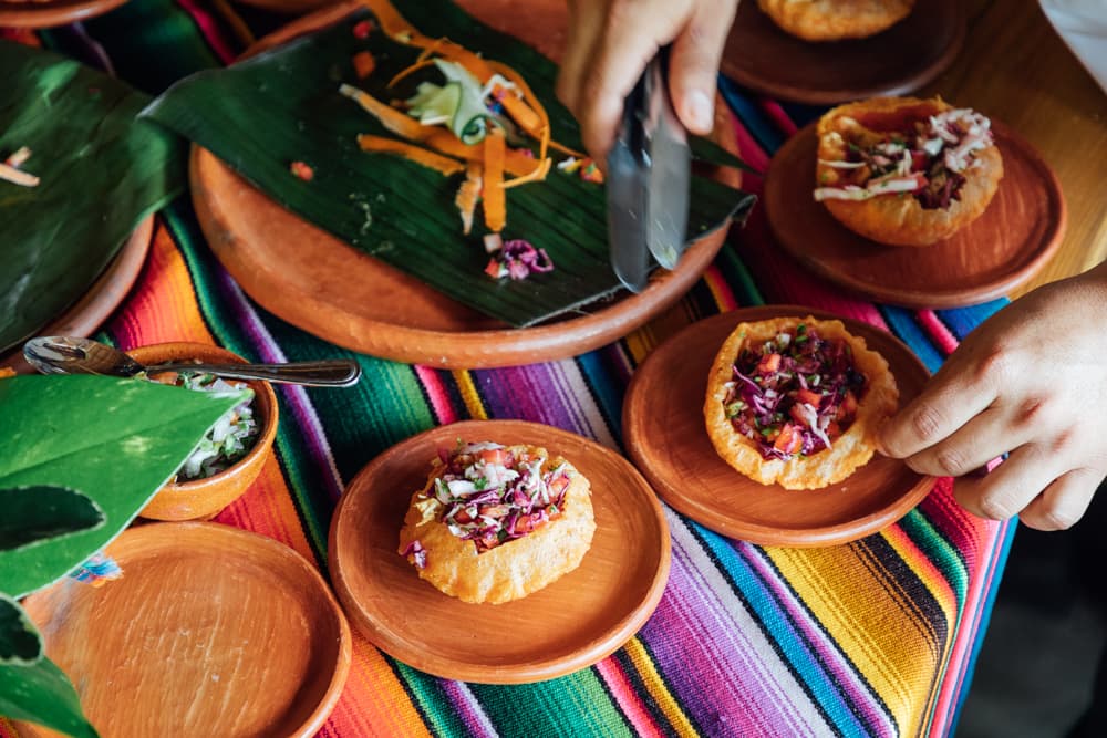 A hand prepares colorful food on wooden plates atop a vibrant woven tablecloth.
