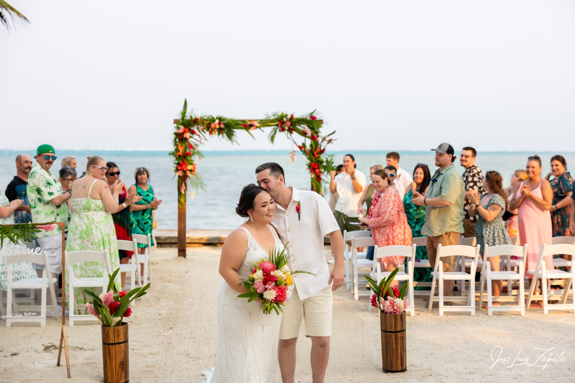A bride and groom kiss joyfully at their beachside wedding, surrounded by smiling guests in a tropical setting.