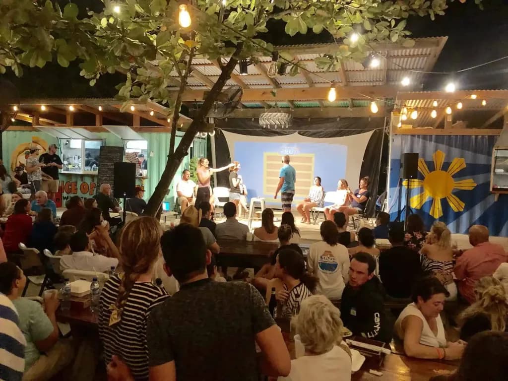 A lively outdoor event with a performance happening on stage, surrounded by an audience seated at tables under string lights.